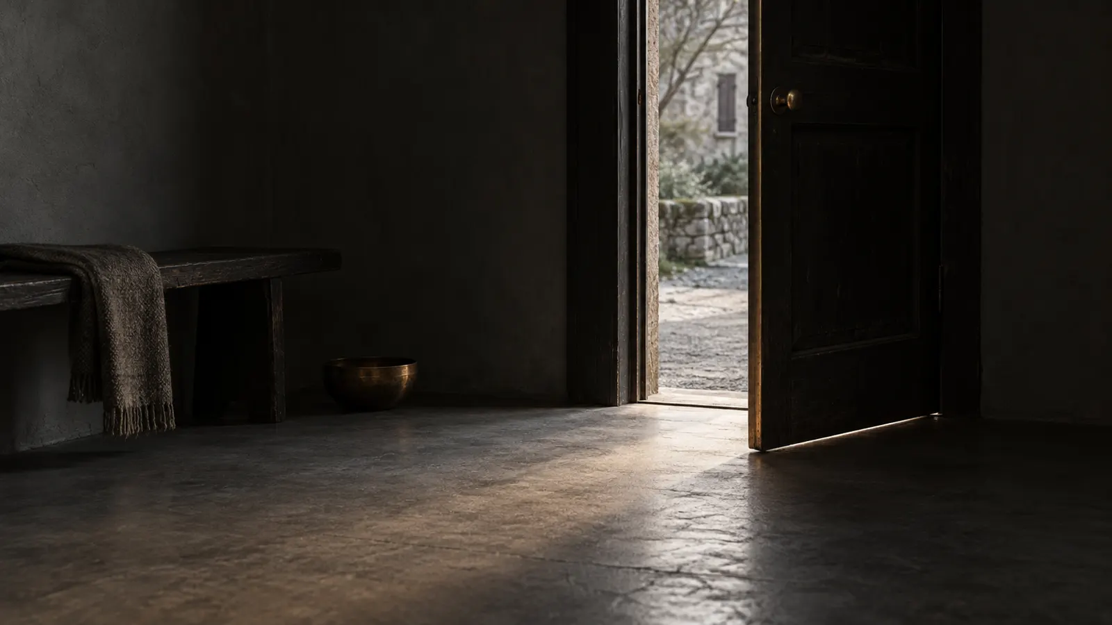 An ajar door in a dark room lets in soft daylight over a gray floor and a slightly bronze-colored threshold.
